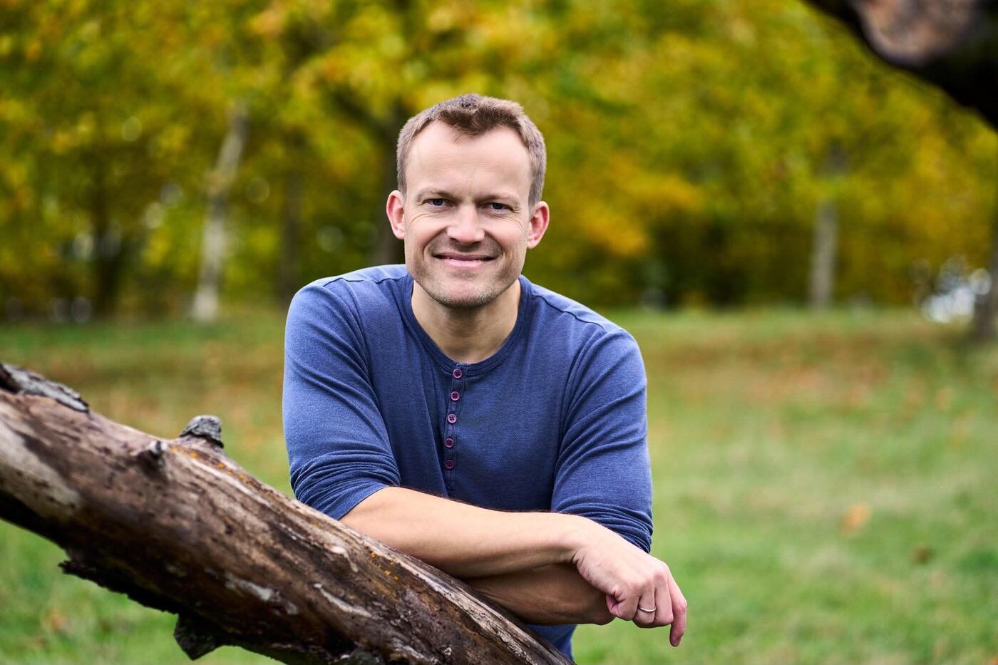 Thomas Jørgensen i blå trøje smiler, lænet op ad en træstamme i en park